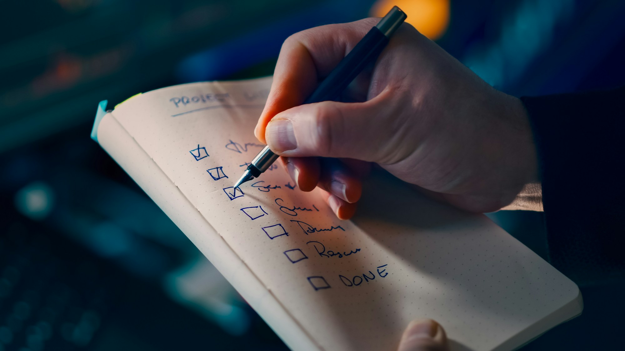 Hand marking a check beside a handwritten project list in a small notebook