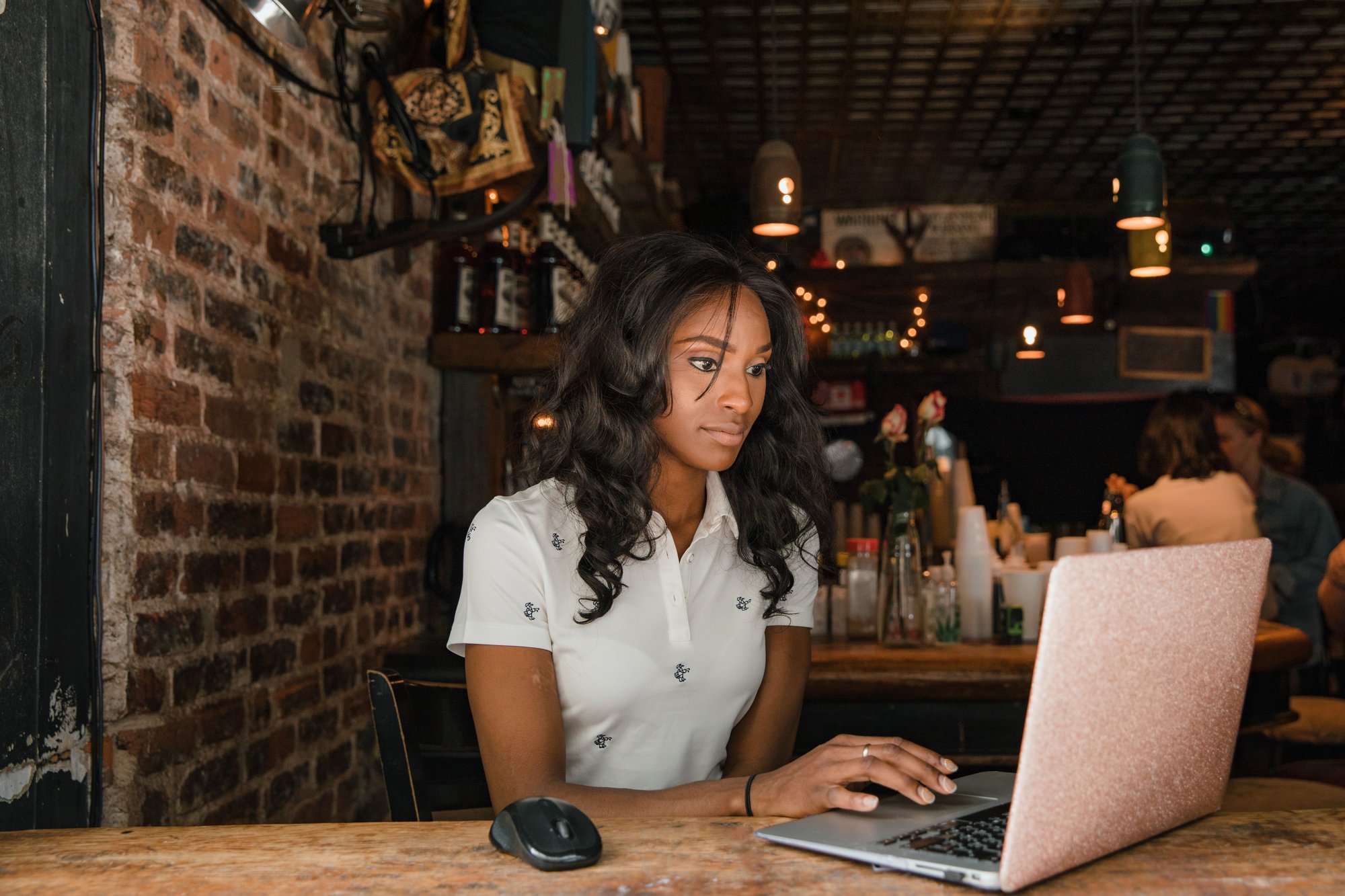 A woman in a white polo types on a rose-gold laptop at a wooden cafe counter, the screen turned away