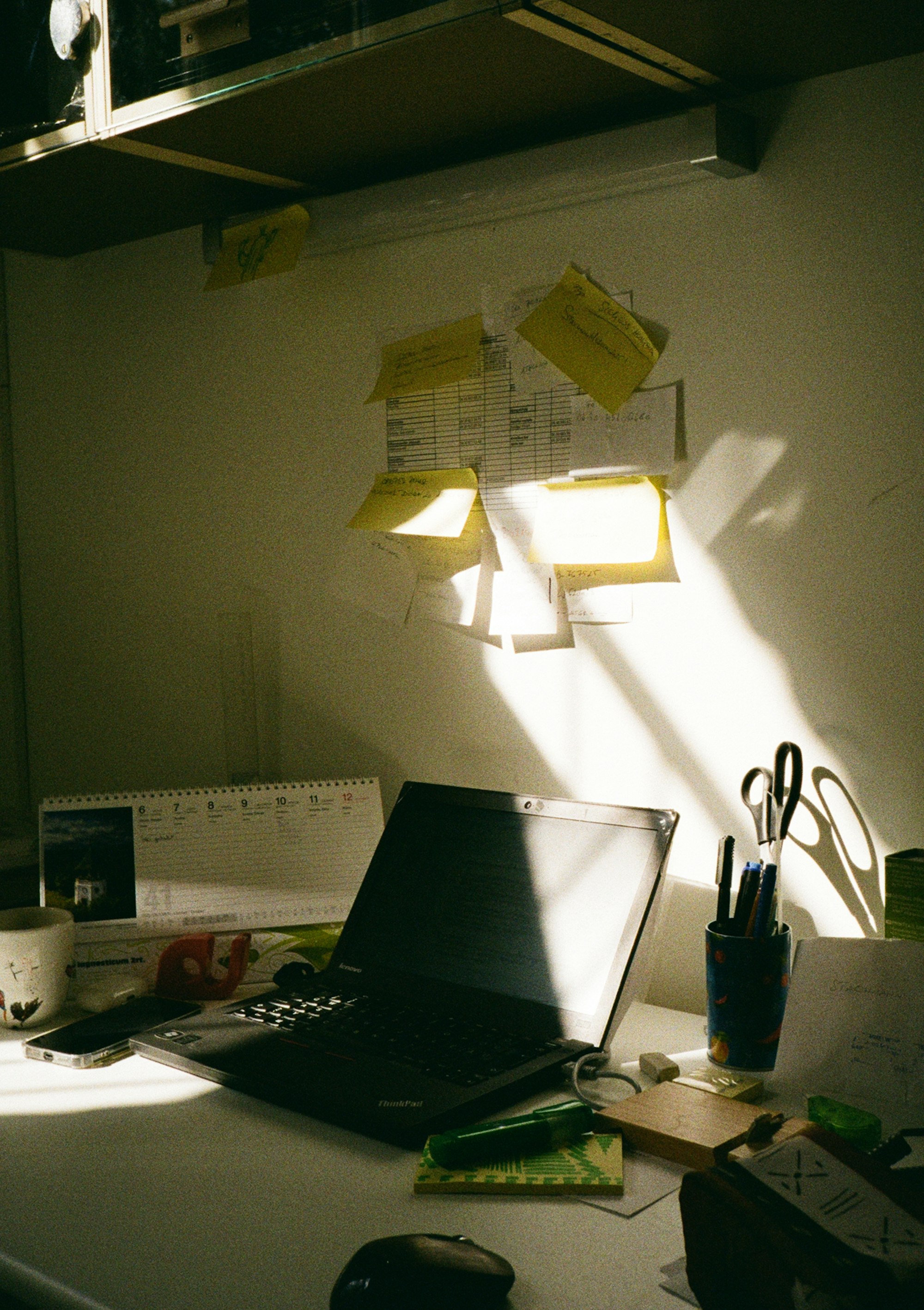 Laptop on a white desk under sticky notes and a printed grid taped to the wall in hard sunlight