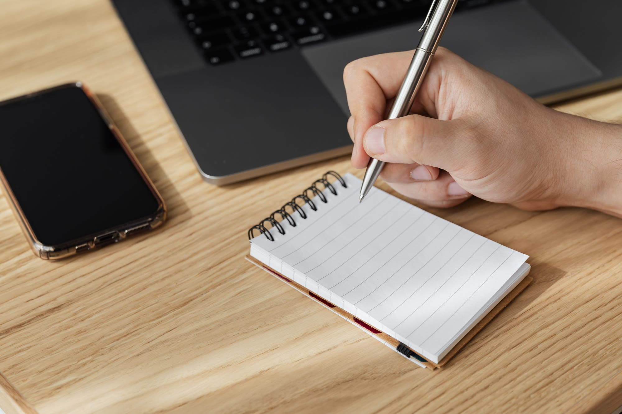 A hand writing in a blank notepad beside a smartphone and a laptop on a wooden desk