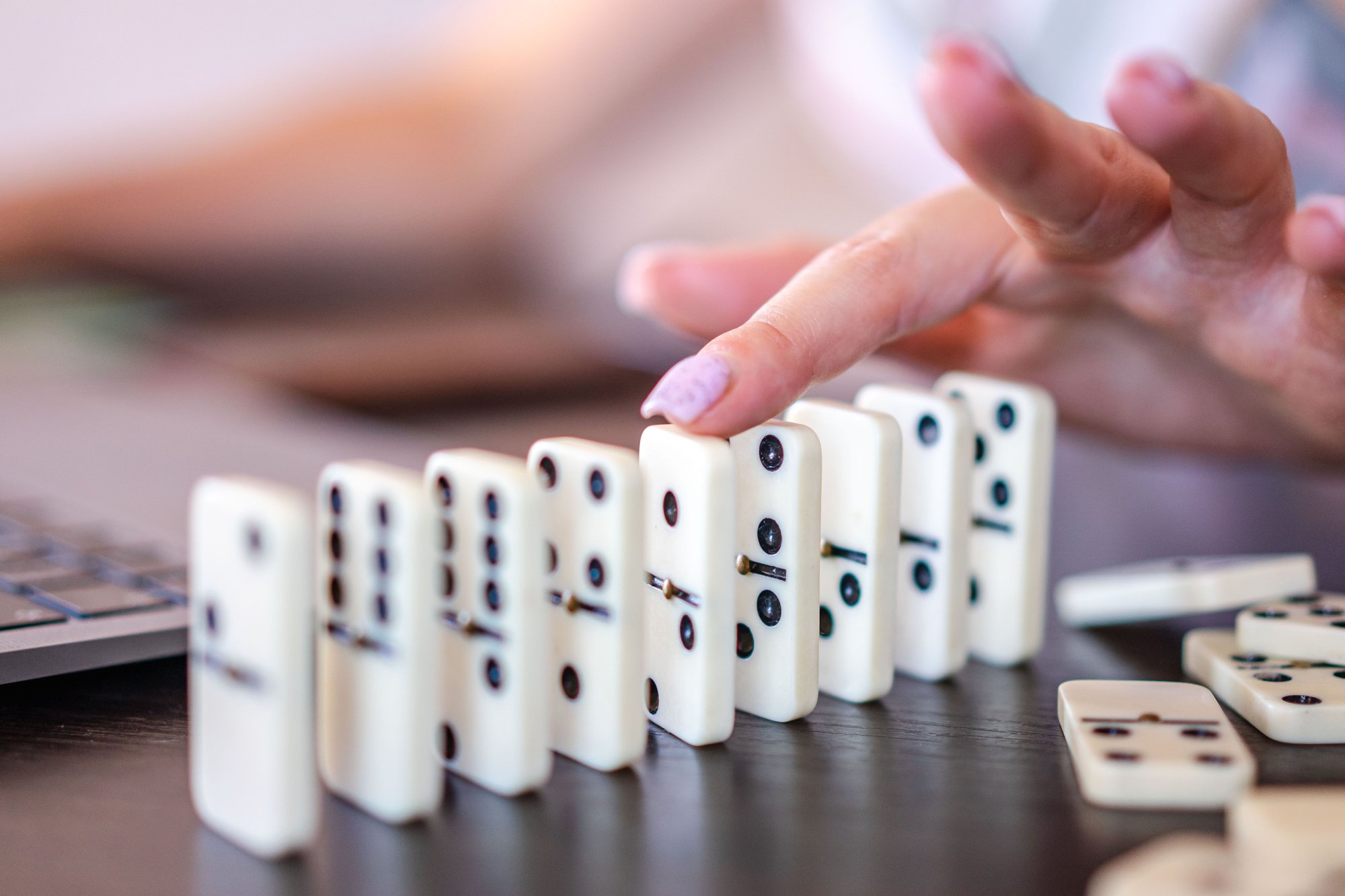 A hand tips the first domino in a row on a dark desk, with a laptop blurred behind it