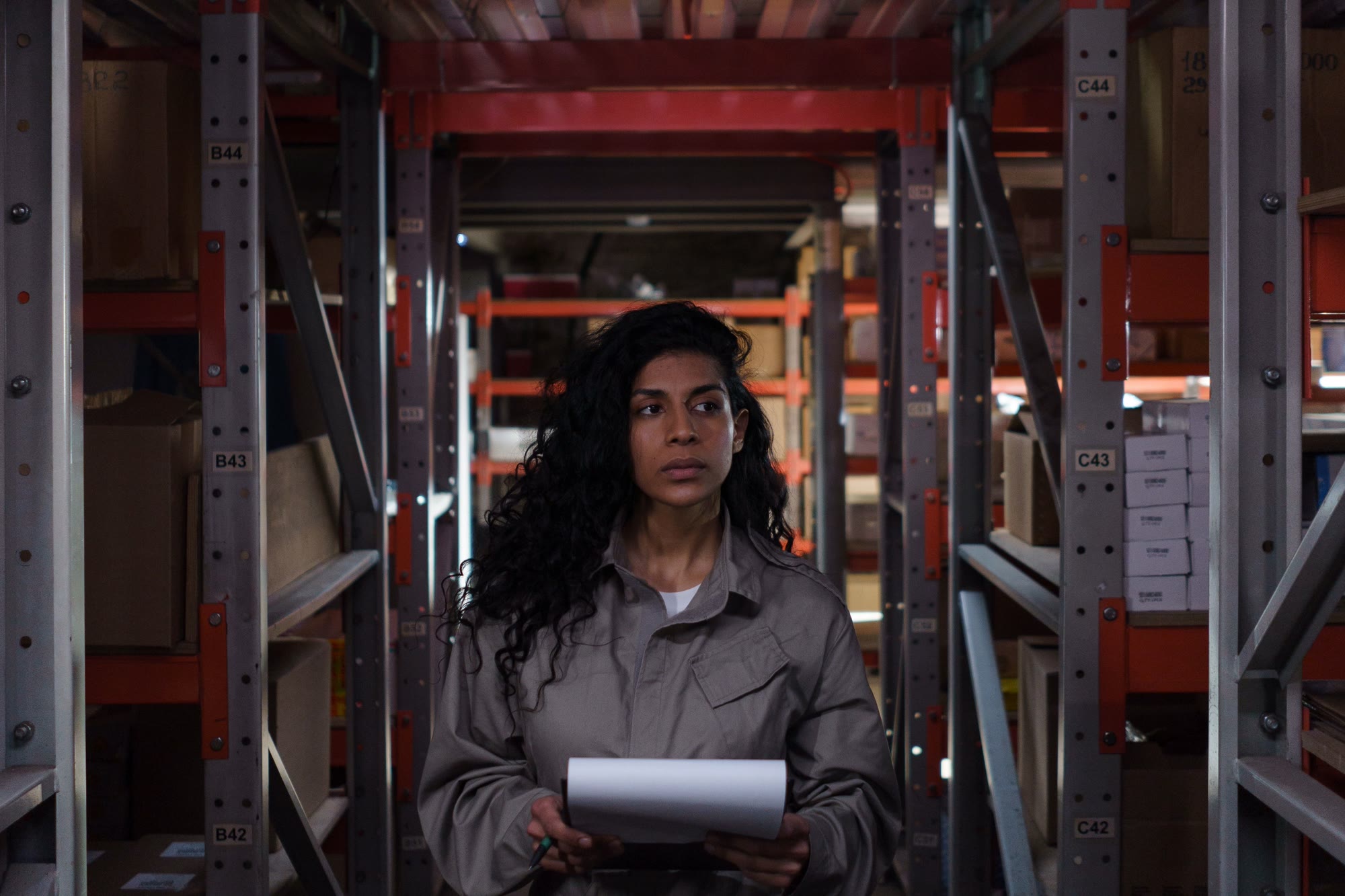 A warehouse worker in a gray uniform stands in a storage aisle holding a blank sheet of paper