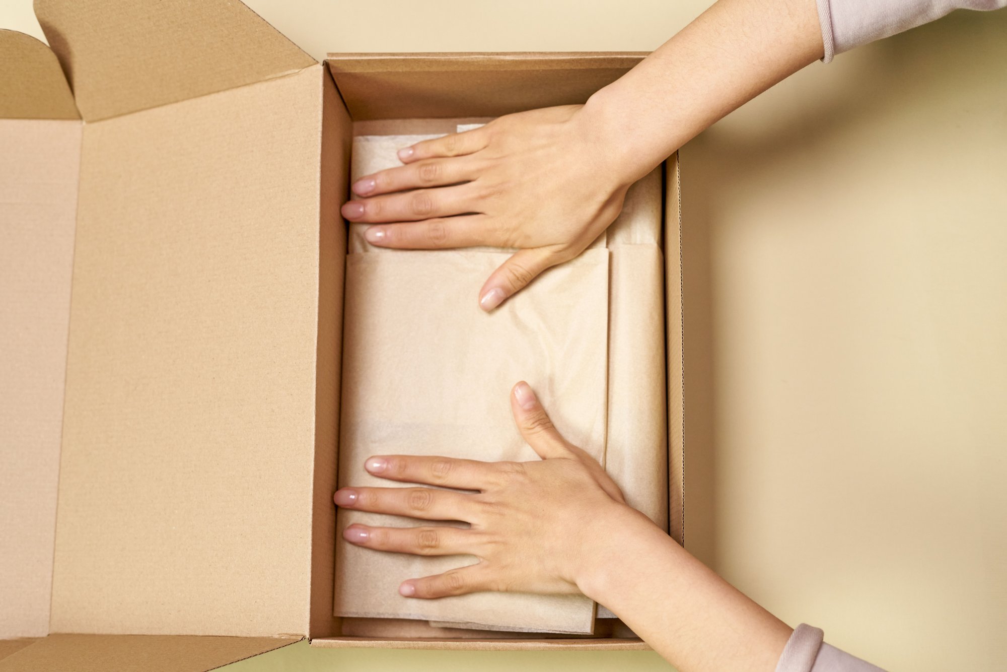 Hands press tissue-wrapped items into an open cardboard box on a pale table