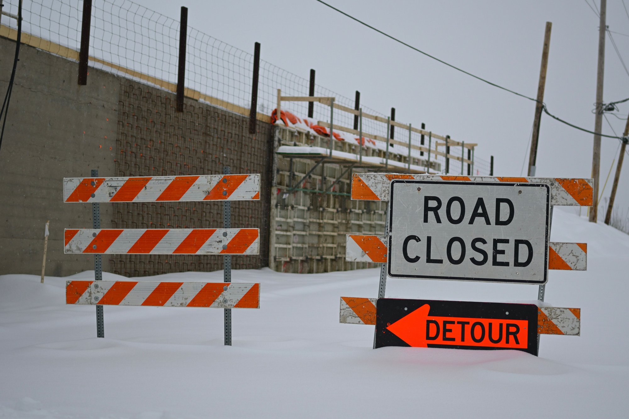 A ROAD CLOSED sign above an orange DETOUR arrow on snowy construction barricades