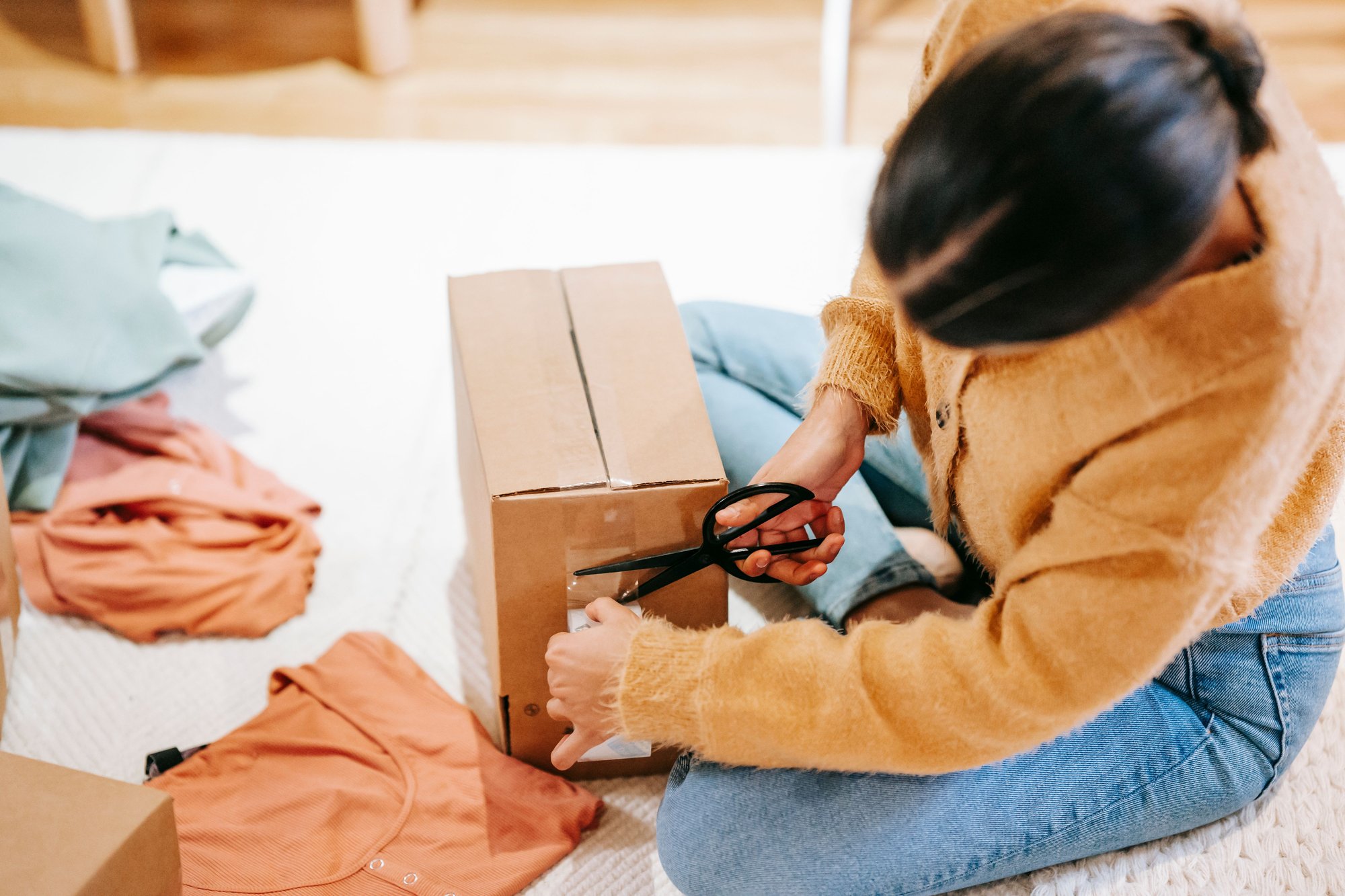 A woman in a mustard sweater sits on the floor sealing a cardboard box with tape, clothes laid out beside her