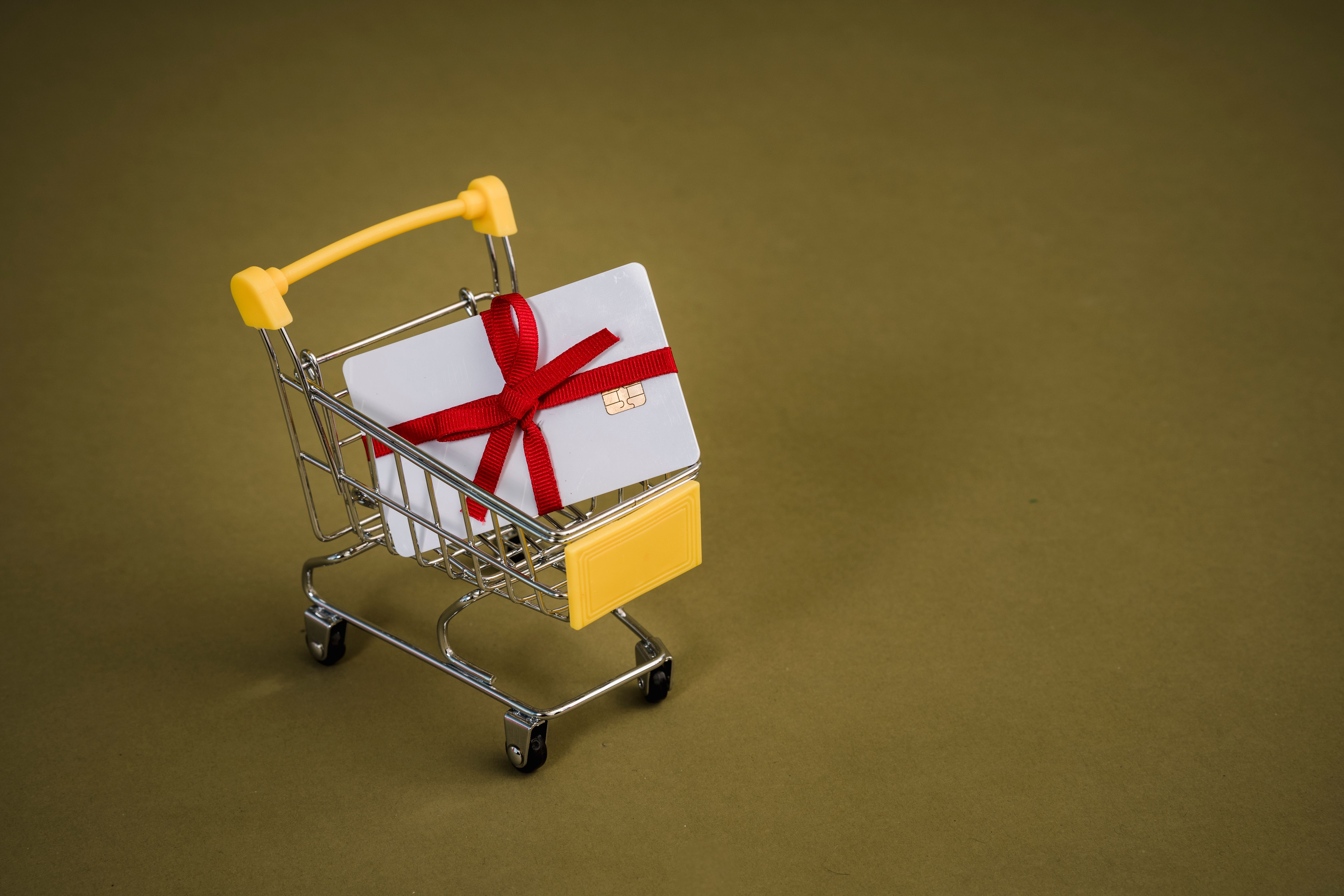 Mini shopping cart holding a white gift card tied with a red ribbon on an olive backdrop